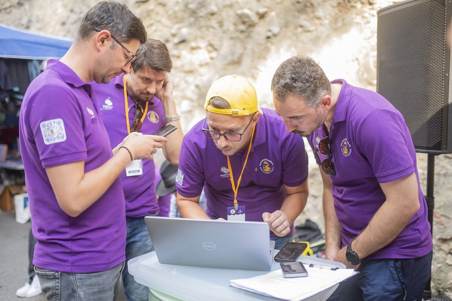 Patru membri RT6 în tricouri mov, aplecați peste un laptop și telefoane la standul cursei.